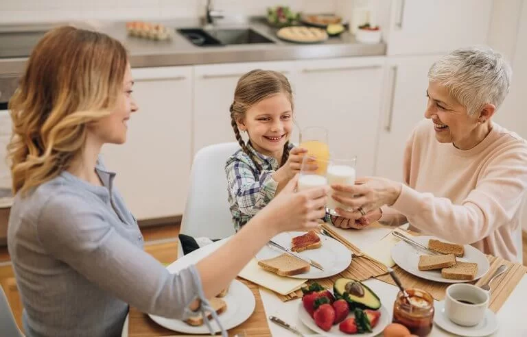 Café da manhã saudável em família: criança, adulta e idosa brindando com leite e suco