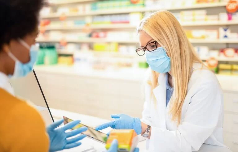 Farmacêutica loura de jaleco branco, óculos e máscara atendendo na farmácia uma paciente