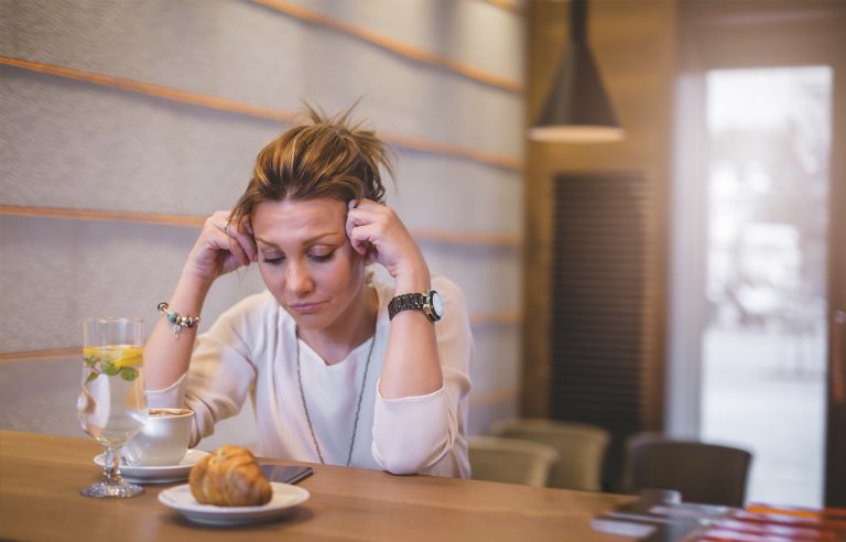 Mulher com expressão de preocupação em mesa de bistrô com comida na frente, bebida e café