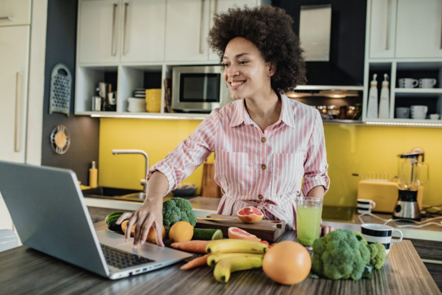 Mulher na cozinha com frutas, legumes, vegetais e com notebook para fazer receita saudável